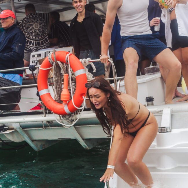 A woman in a bikini reaches for a floating tube in the water while leaning off a boat. A man in swim shorts holds her arm to help. Several people watch from the boat, and bright pool floats are visible in the background.