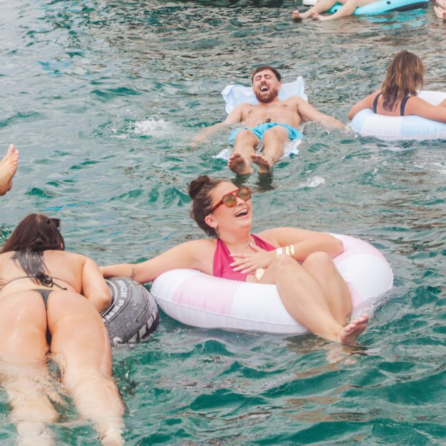 People relaxing on inflatable pool floats in the water, laughing and enjoying themselves on a sunny day. Some are lying on their backs, while others are lounging on colorful tubes. The scene is lively and playful.