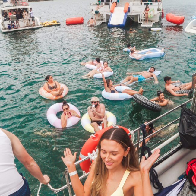 A group of people relax on pool floats and swim in a harbor, with several boats anchored nearby. In the foreground, a woman in a yellow bikini stands on a boat deck, smiling and raising her hands. The scene is lively and festive.