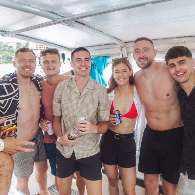 A group of six men and one woman smiling and posing together on a boat. Some hold drinks, and the woman is wearing a red bikini top. The setting looks casual and relaxed, with water and trees visible outside.