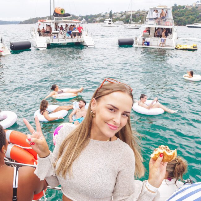 A woman smiles and makes a peace sign while holding food on a boat. Behind her, people relax on inflatables in the water near several boats during a lively gathering on a sunny day.
