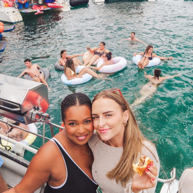 Two women smiling on a boat, one holding food. Behind them, people relax and swim in the water and lounge on inflatable rings, with several boats anchored nearby on a sunny day.