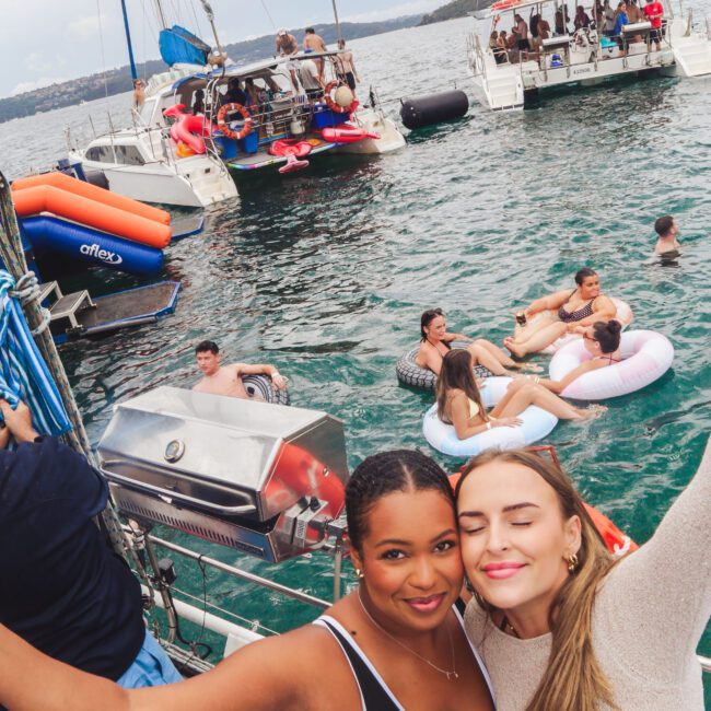 Two women smile and pose for a selfie on a boat, with people swimming and relaxing on floats in the water. Other boats with partygoers are anchored nearby under a cloudy sky.