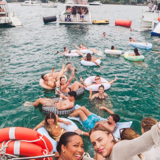 A group of people relax and socialize on floaties in the water near several boats on a cloudy day. Two women smile and pose for the camera in the foreground, with life preservers visible on the boat.