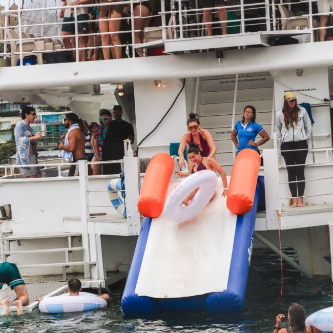 People enjoy a boat party; a woman slides down an inflatable slide from the boat into the water, while others watch from the boat deck or float in the water with tubes.