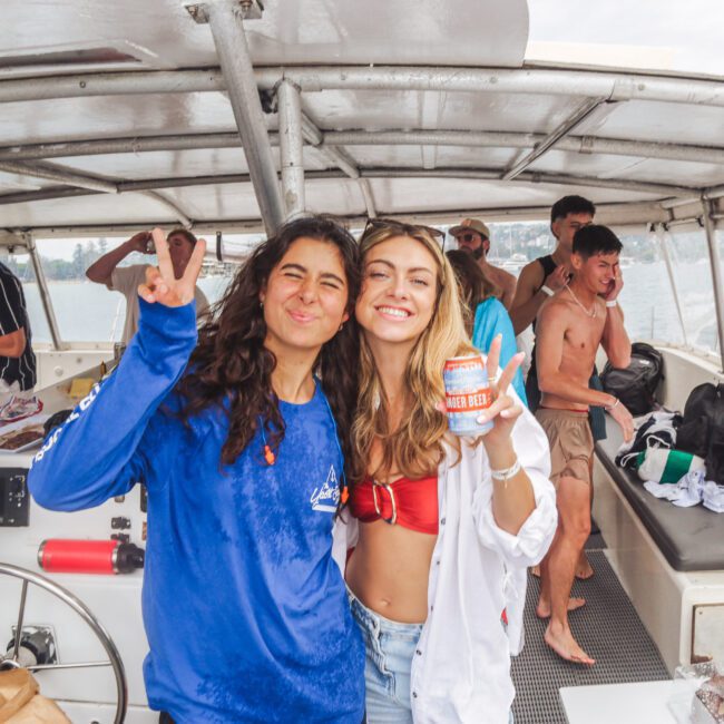 Two smiling women pose on a boat; one makes a peace sign, the other holds a can of beer. Several people are in the background, laughing and enjoying themselves. The boat is on the water, with buildings visible in the distance.