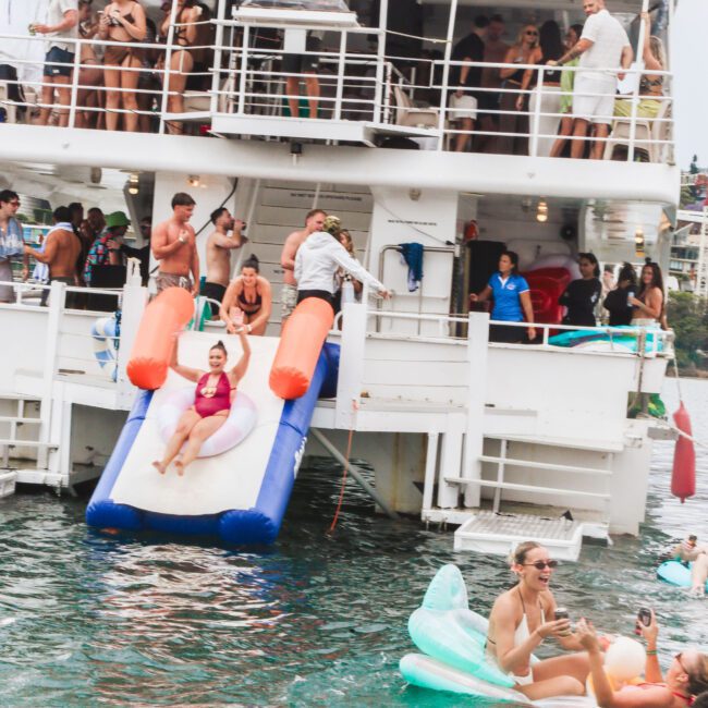 A crowded boat party with people on the deck and in the water. A girl slides down an inflatable slide into the lake, while others relax on pool floats and watch from the boat. Everyone appears to be enjoying the summer day.
