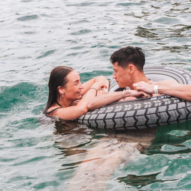 A smiling woman in the water holds onto a flotation ring with a man sitting inside. They look at each other and smile, surrounded by rippling green water.
