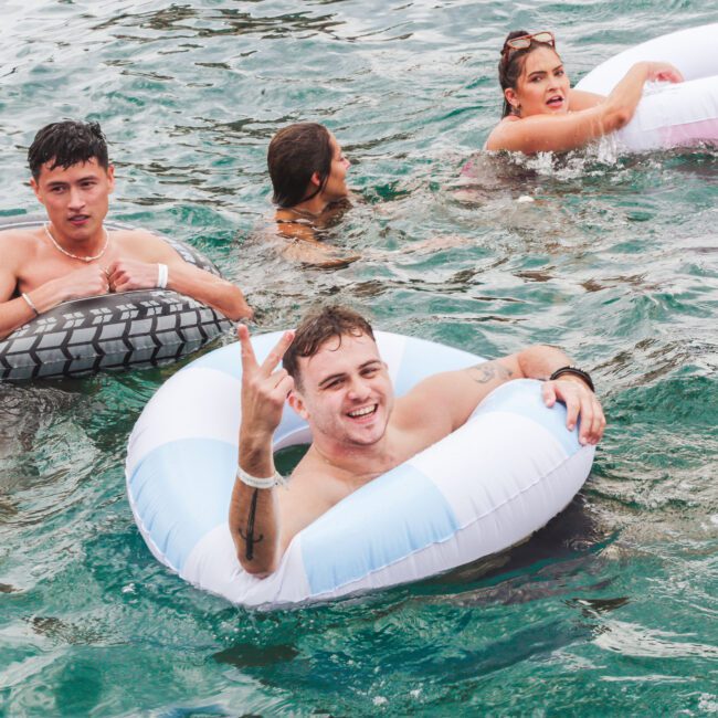 Four young adults enjoy swimming in clear blue water, three of them floating on inflatable rings. One man in front smiles and flashes a hand sign toward the camera. The group looks relaxed and playful.