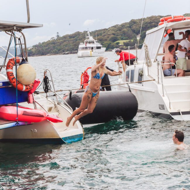 A woman in a blue bikini jumps from a boat into the water near another person swimming, while a group of people socialize on a nearby boat. Several boats are anchored, with a forested shoreline in the background.