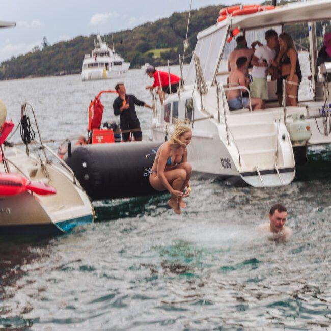 A woman in a swimsuit jumps into the water from a boat while people watch from nearby boats; a man is swimming below her. The scene is lively and set on a sunny day with distant hills in the background.