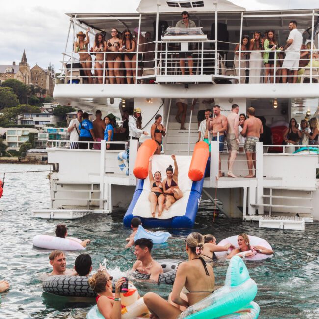 A group of people are partying on a boat and swimming in the water with inflatables. Some are on the upper deck, and others use a slide to enter the water. The scene looks festive and lively.