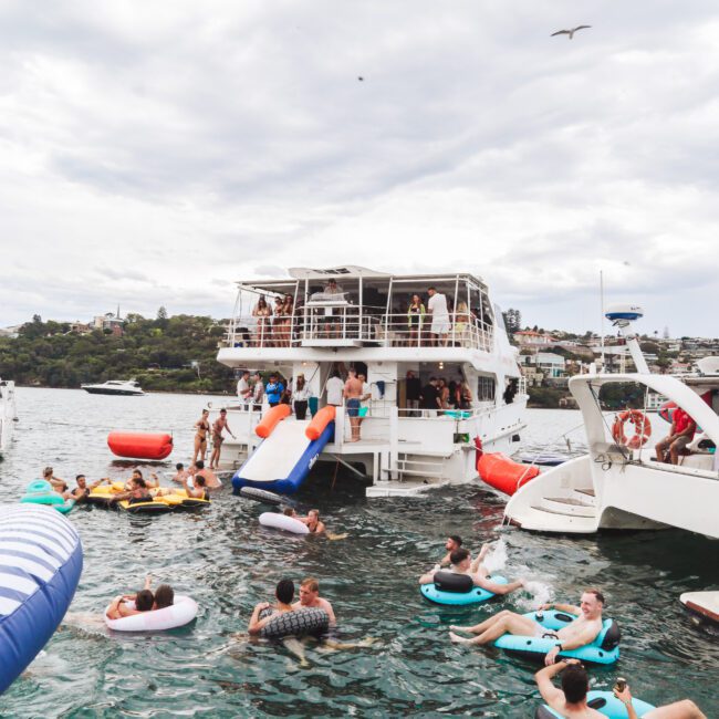 People relax and socialize on inflatables and swim near docked boats in a lively harbor scene. Some are on a large yacht, while others float on the water. The sky is overcast and birds fly above.