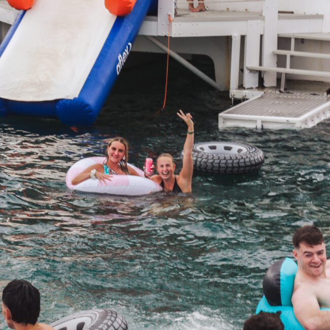 Two women in float rings smile and wave at the camera while enjoying the water near a boat with a slide. Other people with tire-shaped float rings swim nearby. The scene is lively and festive.