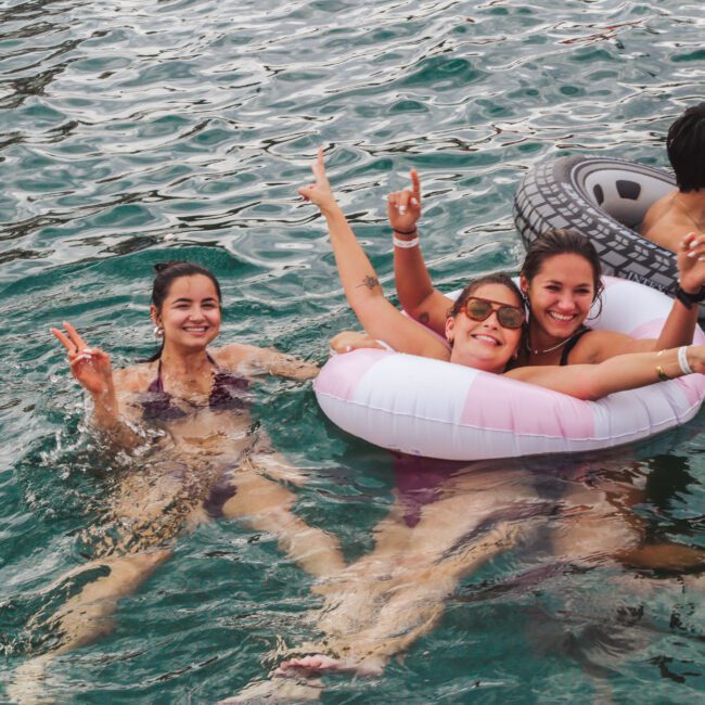 Three young women smile and pose for the camera while floating in water, with two of them sharing a pink inflatable ring. A man on a gray tire float faces away from the camera. Everyone appears to be enjoying a fun time outdoors.