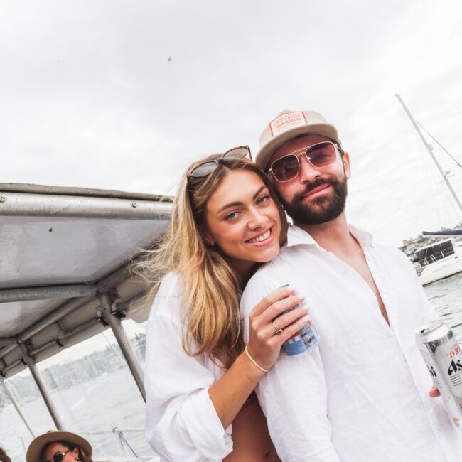 A smiling woman and a bearded man in sunglasses and a cap pose together on a boat. Both hold drinks, with water and other boats visible in the background.