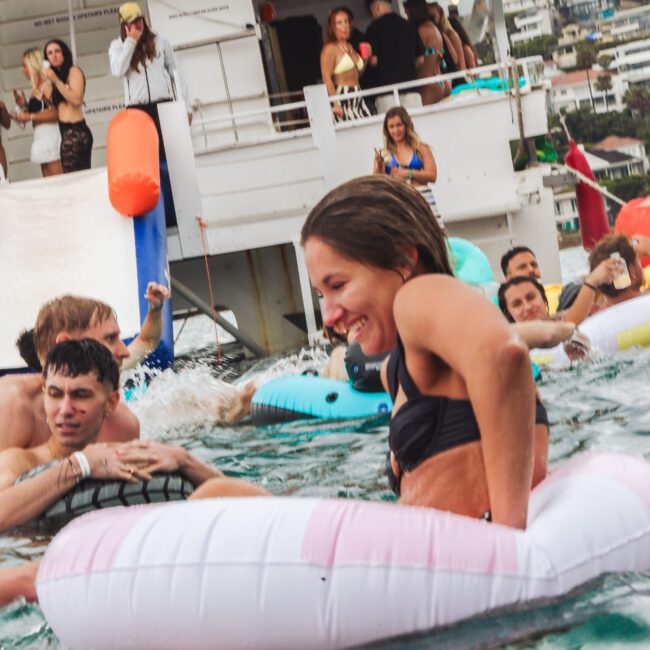 A woman in a black bikini smiles while sitting on a pink and white inflatable ring in the water, surrounded by people swimming and partying near a large boat. Other partygoers watch from the deck above.