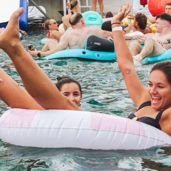 Two young women laugh and play in a pool, one sitting in a white and pink inflatable ring with her legs up, surrounded by other people relaxing on floaties in the water.