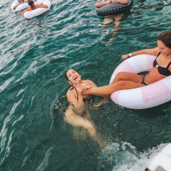 Two women laugh and play in clear blue water, one sitting in a pink and white inflatable ring while the other holds her feet and floats nearby. Other people on inflatables are visible in the background.