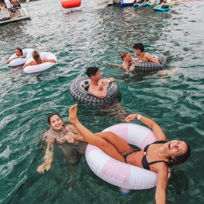 People are relaxing and playing in a lake, some floating on inflatable tubes. Two women smile at the camera, one inside a pink tube, and others enjoy the water nearby. Boats and more people are visible in the background.