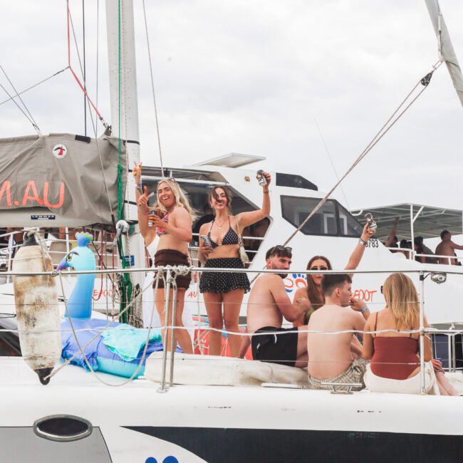 A group of people in swimwear smile and raise drinks while relaxing on the deck of a catamaran, docked near another boat on a cloudy day.