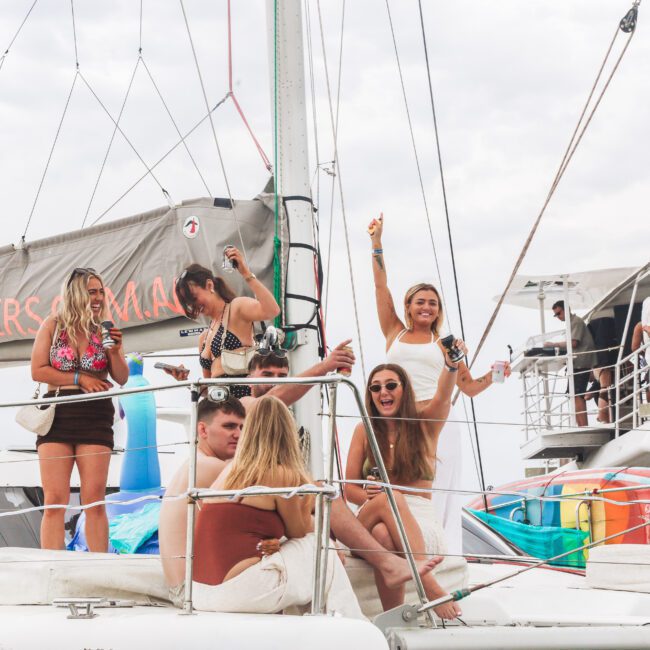 A group of young adults relax and celebrate on the deck of a sailboat, laughing, holding drinks, and enjoying a sunny day. The boat is anchored, with colorful inflatables and a second deck visible in the background.