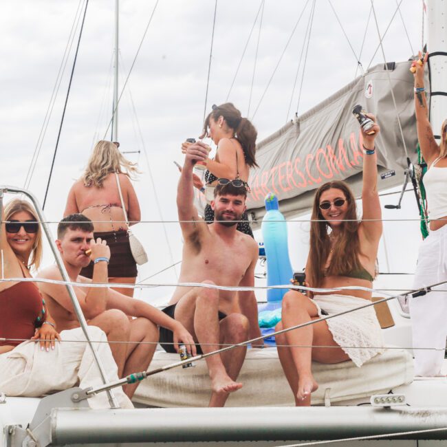Six people relax and raise drinks while sitting on the deck of a sailboat, smiling and enjoying themselves. Some are wearing sunglasses, swimsuits, and summery clothes. A banner is visible in the background.