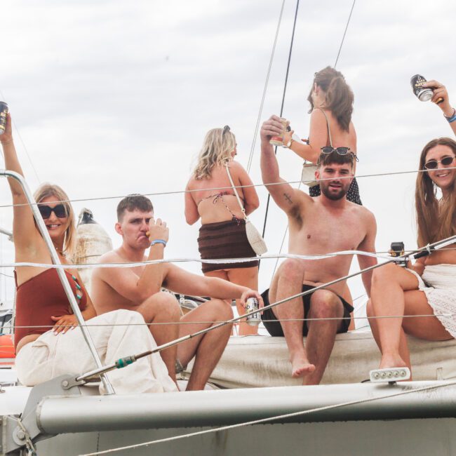 Four young adults sit and smile on the deck of a boat, raising drinks in a celebratory toast. Two women and two men are in the foreground, with three more people visible in the background. The sky appears overcast.