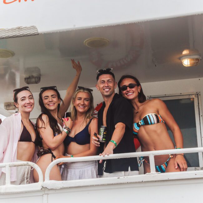 Five people in swimwear smile and pose together on the deck of a boat, some holding drinks. They appear to be enjoying a sunny day, with bright expressions and casual summer outfits.