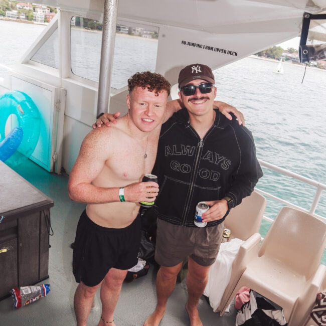 Two smiling men stand on a boat. One is shirtless, wearing black swim trunks, and holding a drink. The other wears a cap, sunglasses, a hoodie, and shorts, also holding a drink. Water and shoreline are visible behind them.