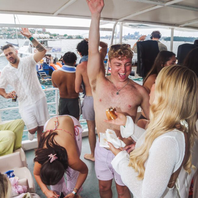 A group of young adults enjoy a lively boat party. A shirtless man with a raised arm smiles at a woman in a white sheer dress as others socialize, eat, and take photos near water with boats in the background.