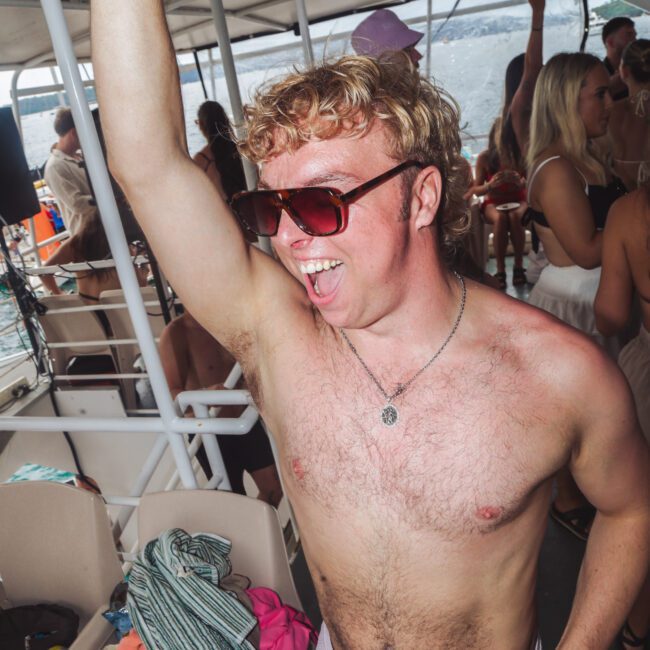 A young man with curly blond hair, sunglasses, and no shirt enthusiastically dances on a boat crowded with people, with his arm raised and mouth open, appearing joyful at a lively party.