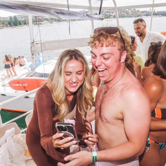 Two people, one in a bikini and cover-up and one shirtless, smile and laugh while looking at a phone during a lively boat party with others nearby and a sailboat visible on the water in the background.