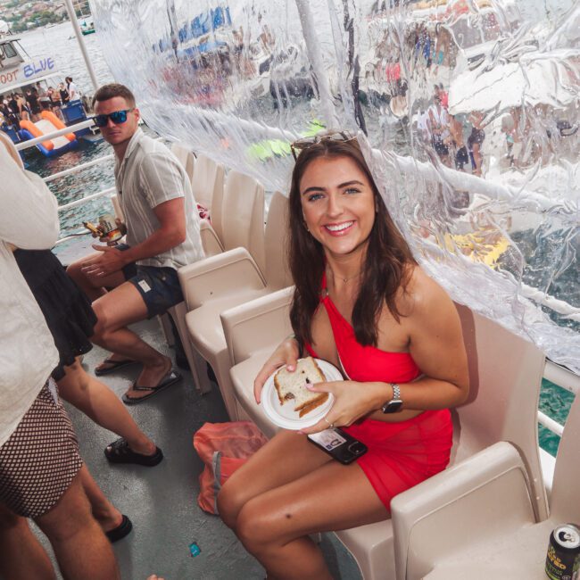 A woman in a red dress sits on a boat seat, smiling at the camera and holding a sandwich on a plate. Other people are seated nearby, and the boat is covered with a transparent plastic sheet. Water and boats are visible outside.