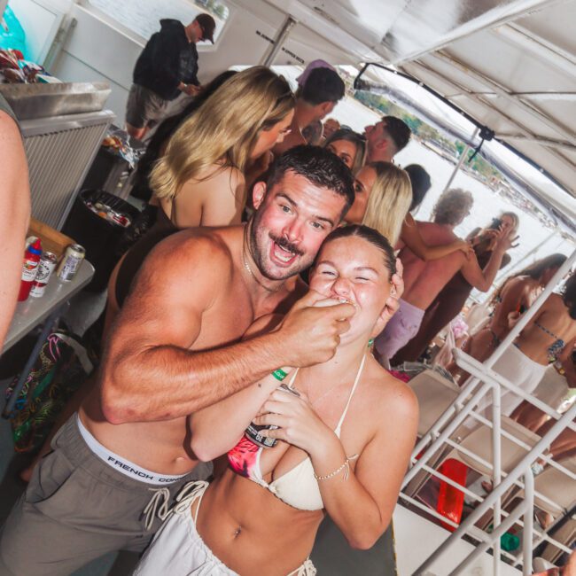 A shirtless man playfully hugs a woman in a white bikini top on a crowded boat. Both are laughing and smiling, surrounded by other people enjoying the party atmosphere.