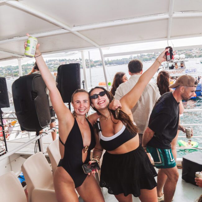 Two young women in swimsuits stand on a boat, smiling with arms raised, holding drinks. Other people lounge or chat around them, and water with boats is visible in the background. It appears to be a lively party scene.