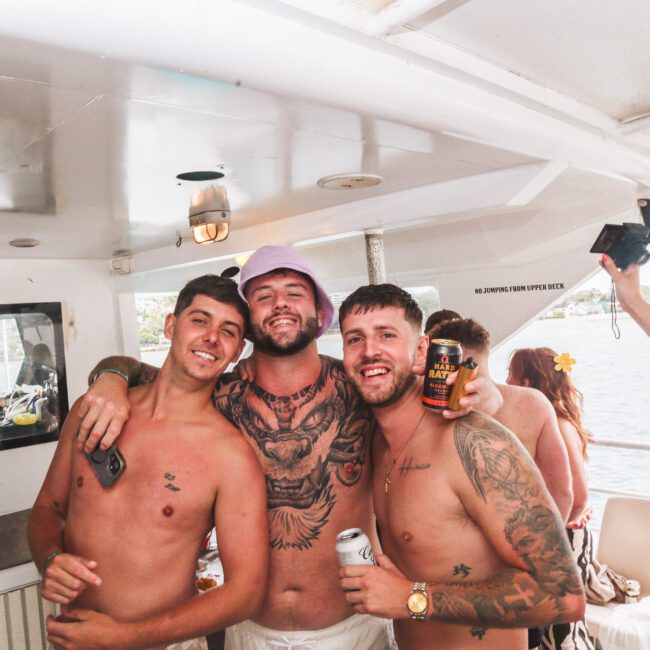 Three shirtless men smiling and posing with drinks on a boat. One wears a purple bucket hat; all have tattoos. Other people are in the background, with water and distant land visible through the windows.