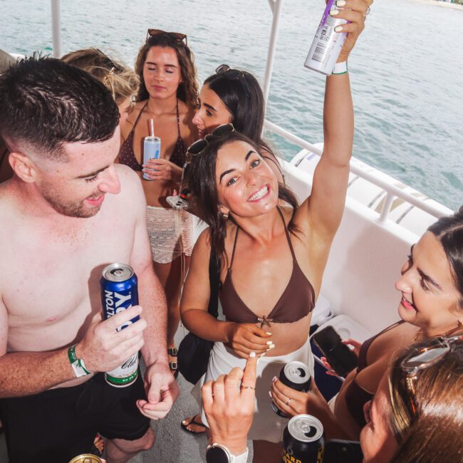 A group of young adults in swimsuits enjoy drinks and smile on a boat, with one woman in the center raising a can in celebration. The water and shoreline are visible in the background.