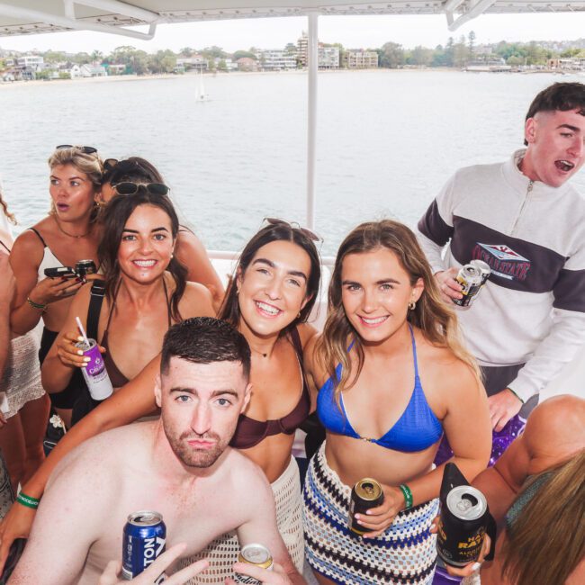 A group of young adults in swimwear and casual clothes smile and hold drinks while posing for a photo on a boat, with water and buildings visible in the background.