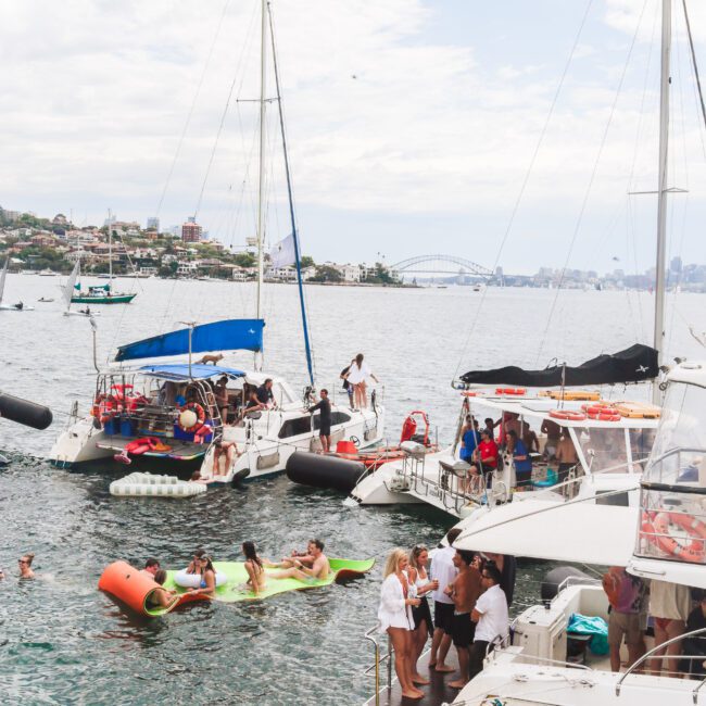 Several boats are anchored close together on a busy harbor, with people socializing on the decks and swimming in the water. Floating inflatables and a distant city skyline are visible in the background.
