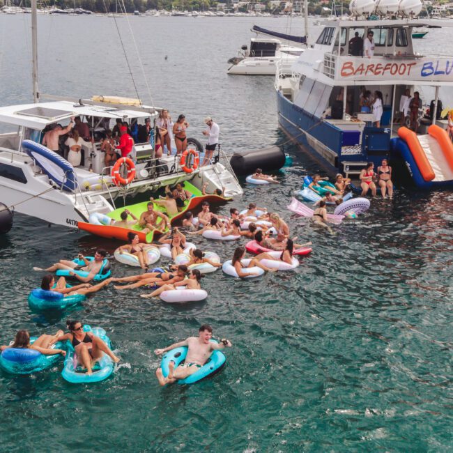A lively group of people float on colorful inflatable tubes near two anchored boats, with some passengers on deck and others swimming in the water on a sunny day. The boats are labeled “BAREFOOT BLUE” and “Barefoot.”.