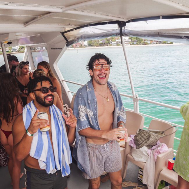 A group of people enjoy a lively boat party by the water. Two men in the foreground smile, wearing swimsuits and holding drinks, one with a towel around his neck. Others socialize in the background near the open deck.