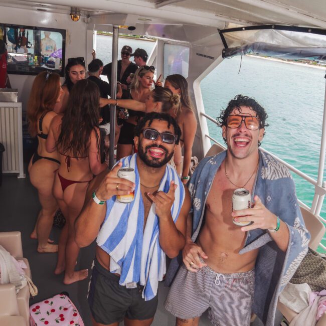 Two smiling men in swim trunks and sunglasses, holding drinks, stand at the front of a boat. Behind them, people in swimsuits are socializing and enjoying the sunny weather near the water.