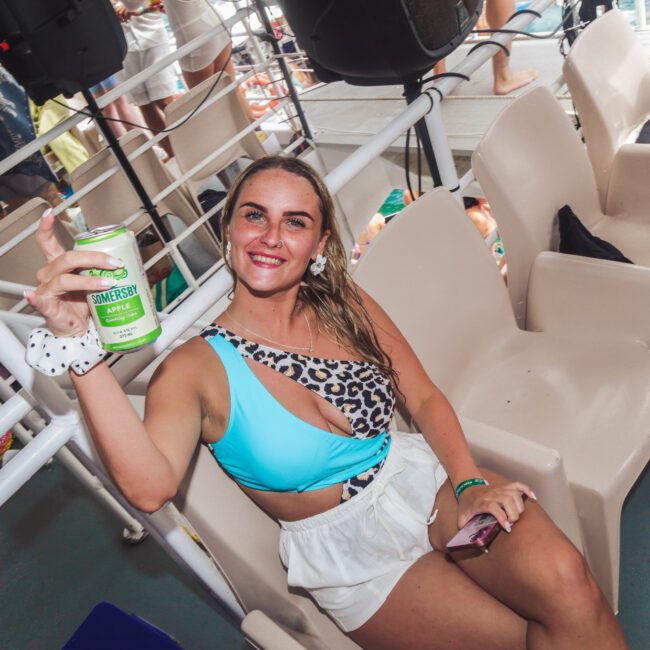 A smiling woman in a stylish swimsuit sits on a boat, holding up a can of drink. Other people are in the background, enjoying the sunny day. The atmosphere is lively and festive.