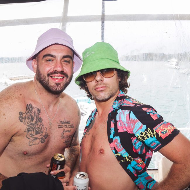 Two men on a boat pose for a photo. Both wear bucket hats, and one is shirtless with tattoos while the other wears an open tropical shirt. They are smiling and holding drinks, with water and boats visible in the background.