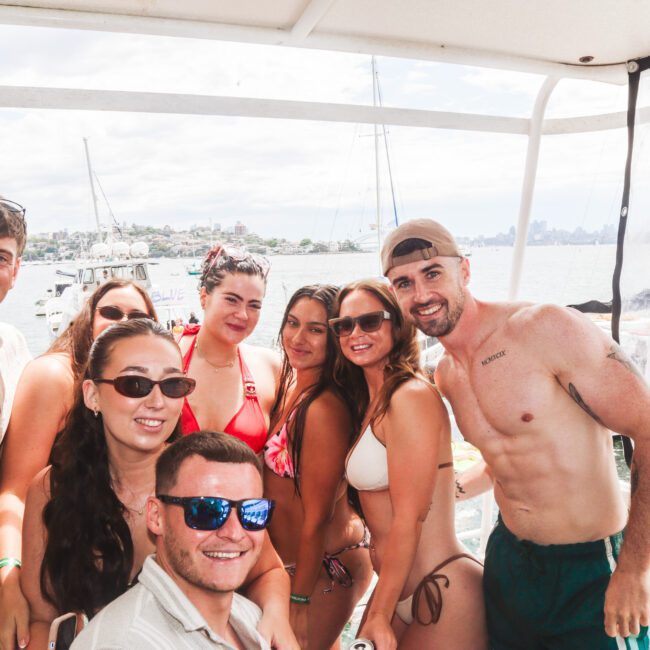 A group of seven smiling friends wearing swimsuits and sunglasses pose together on a boat with water, distant boats, and a city skyline visible in the background on a sunny day.