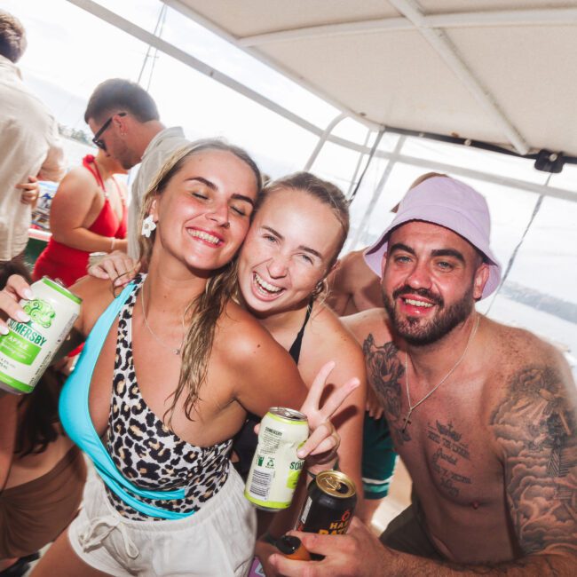 Three smiling people in swimwear pose closely together on a boat, holding drinks. The woman in the middle flashes a peace sign; others are smiling at the camera. More people and water are visible in the background.