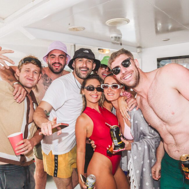 A group of smiling friends in swimsuits and casual summer clothes pose together on a boat, holding drinks and wearing sunglasses and hats. The atmosphere is lively and festive.