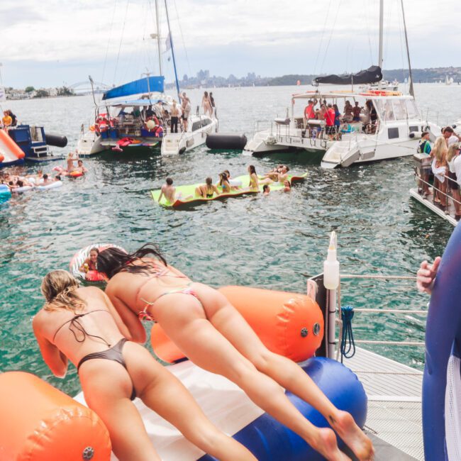 Two women in bikinis dive onto an orange and white inflatable from a boat, surrounded by people swimming, lounging on floats, and other boats on a busy water scene.