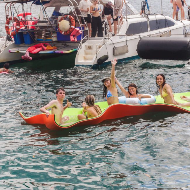 A group of five smiling people relax on a floating mat in the water near two docked boats, enjoying a sunny day. Other people swim and lounge nearby.